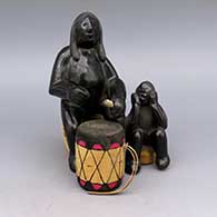 Black figure of man and child, includes wooden log seat, cork seat, wood drumstick, and wood and leather drum