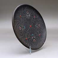 A black micaceous plate with inlaid stones and a sgraffito spider and geometric design, click or tap to see a larger version