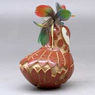 A red and golden micaceous parrot figure decorated with a carved and painted bird element design, plus feathers on the head, click or tap to see a larger version