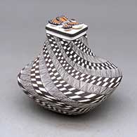 A lidded black on white squarish jar decorated with a spiraling fineline and checkerboard design topped with a polychrome butterfly on the lid
 by Sharon Lewis of Acoma
