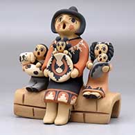 A grandmother storyteller figure sitting on a stone wall with four children, a puppy and a welcome basket, click or tap to see a larger version